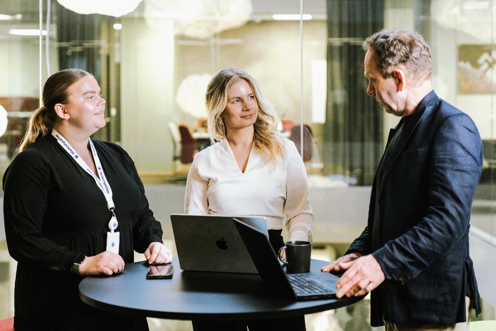 Tre personer som står rundt et bord og snakker og ser på en datamaskin