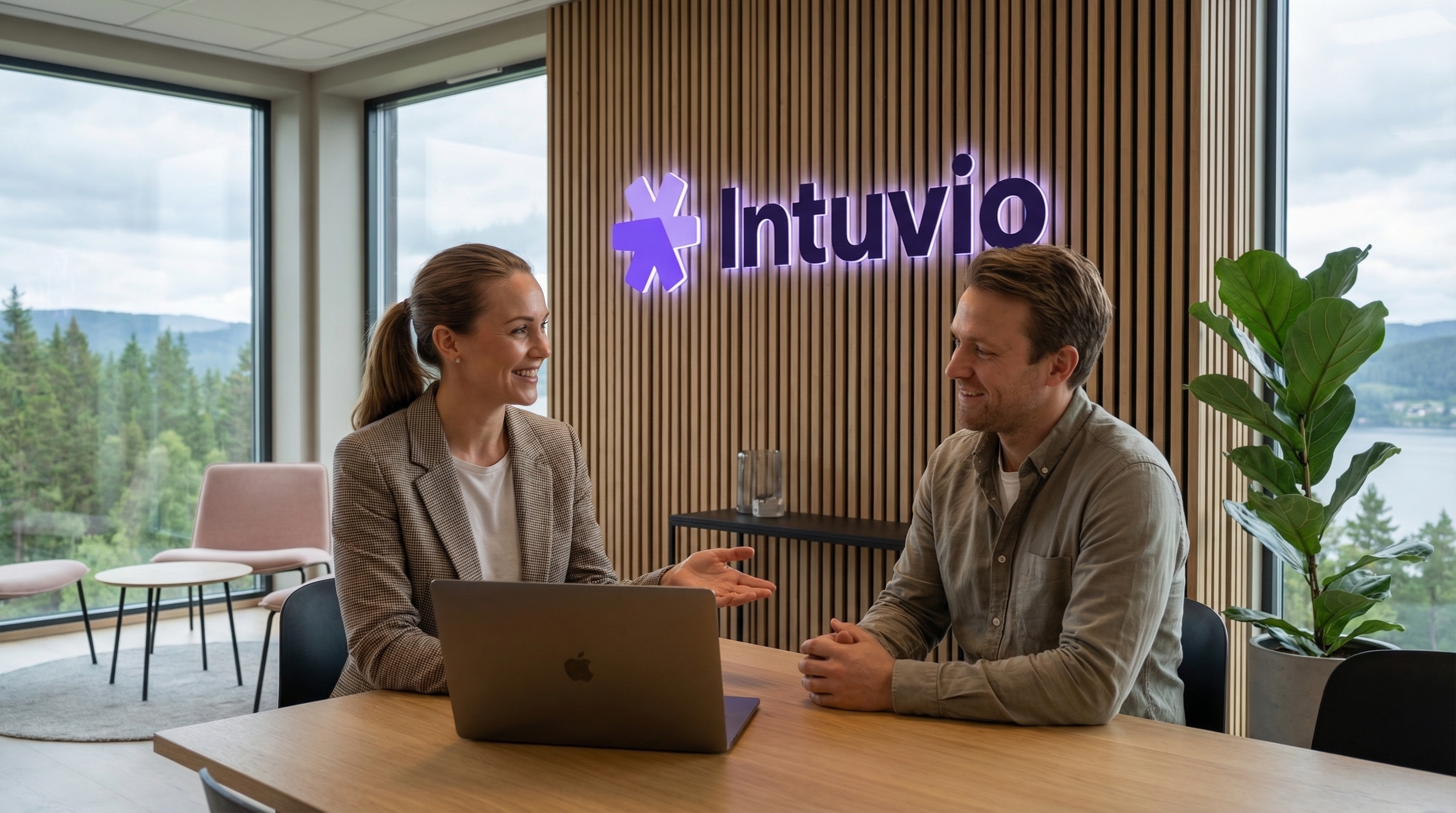 En mann og en kvinne i et positivt forretningsmøte. De smiler og prater over en laptop, med Intuvio-logoen på veggen bak seg.